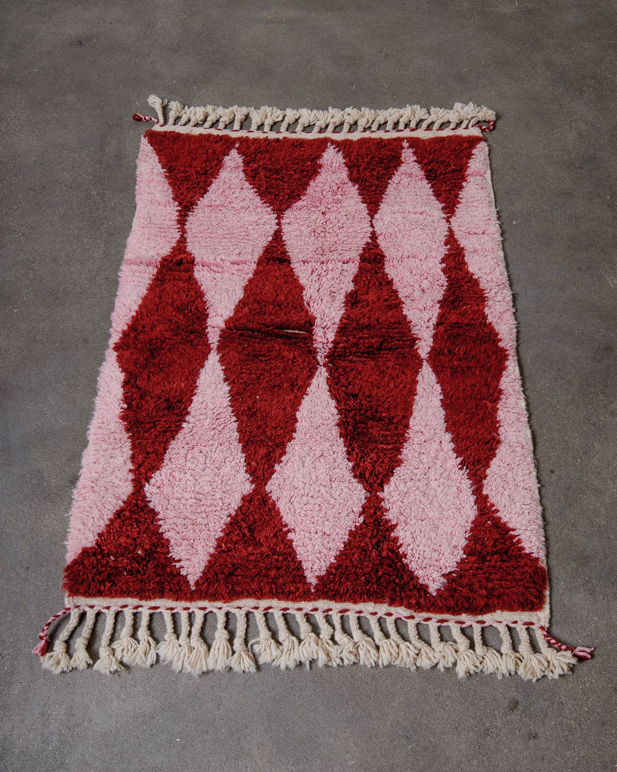 Small Beniourain rug with a harlequin pattern in pink and burgundy wool and with natural wool tassels, laying on concrete floor.